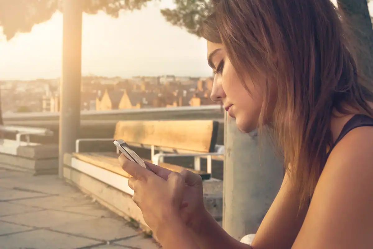 Tânără așezată pe o bancă, ajustând o fotografie pe ecranul telefonului la lumina apusului