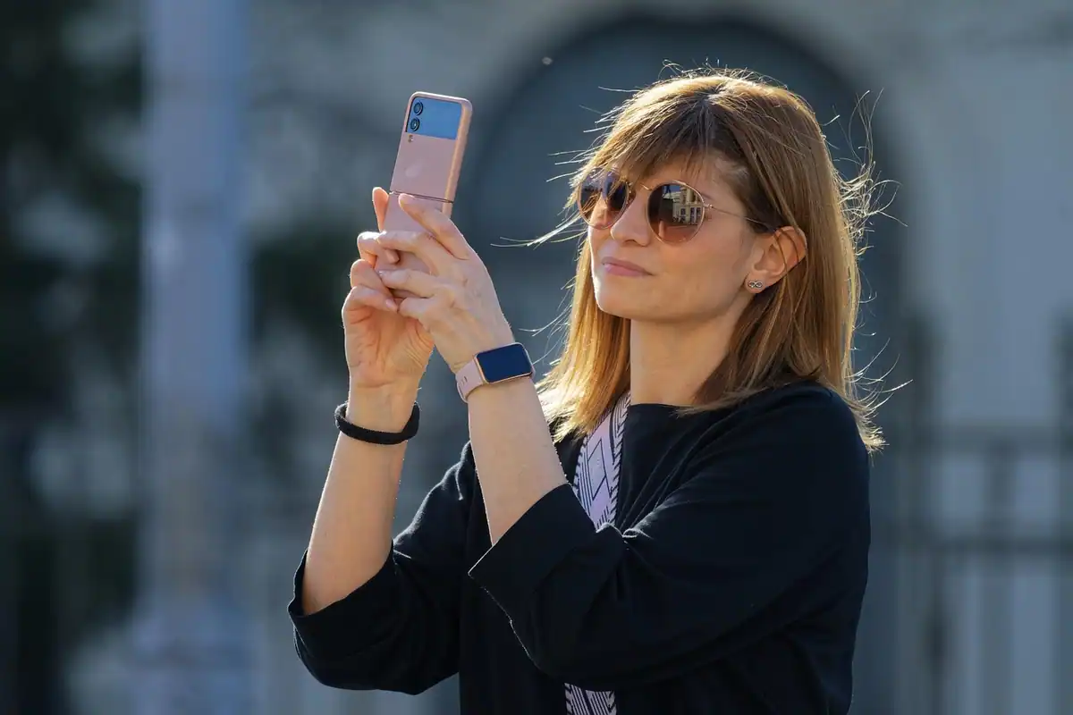Femeie cu ochelari de soare care fotografiază cu telefonul pe stradă, cu clădiri neclare în spate