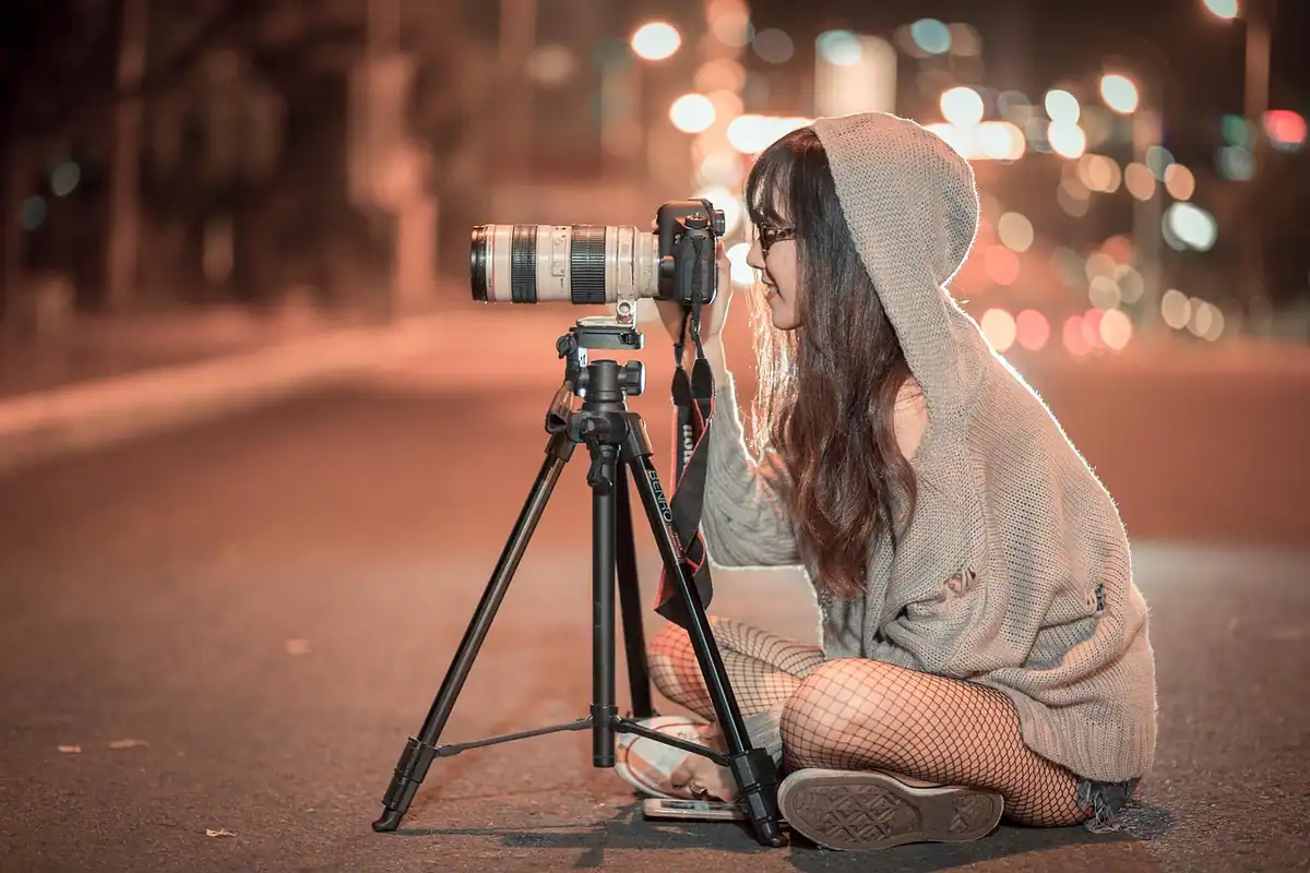Fotografă așezată pe stradă seara, cu aparatul pe trepied, în lumină scăzută