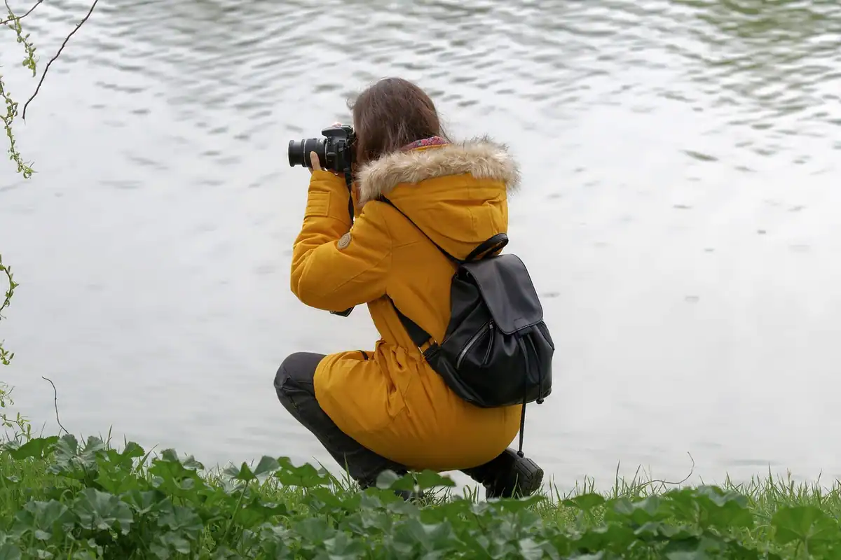 Femeie în geacă galbenă, ghemuită lângă un lac, fotografiază cu aparatul în mod ales manual