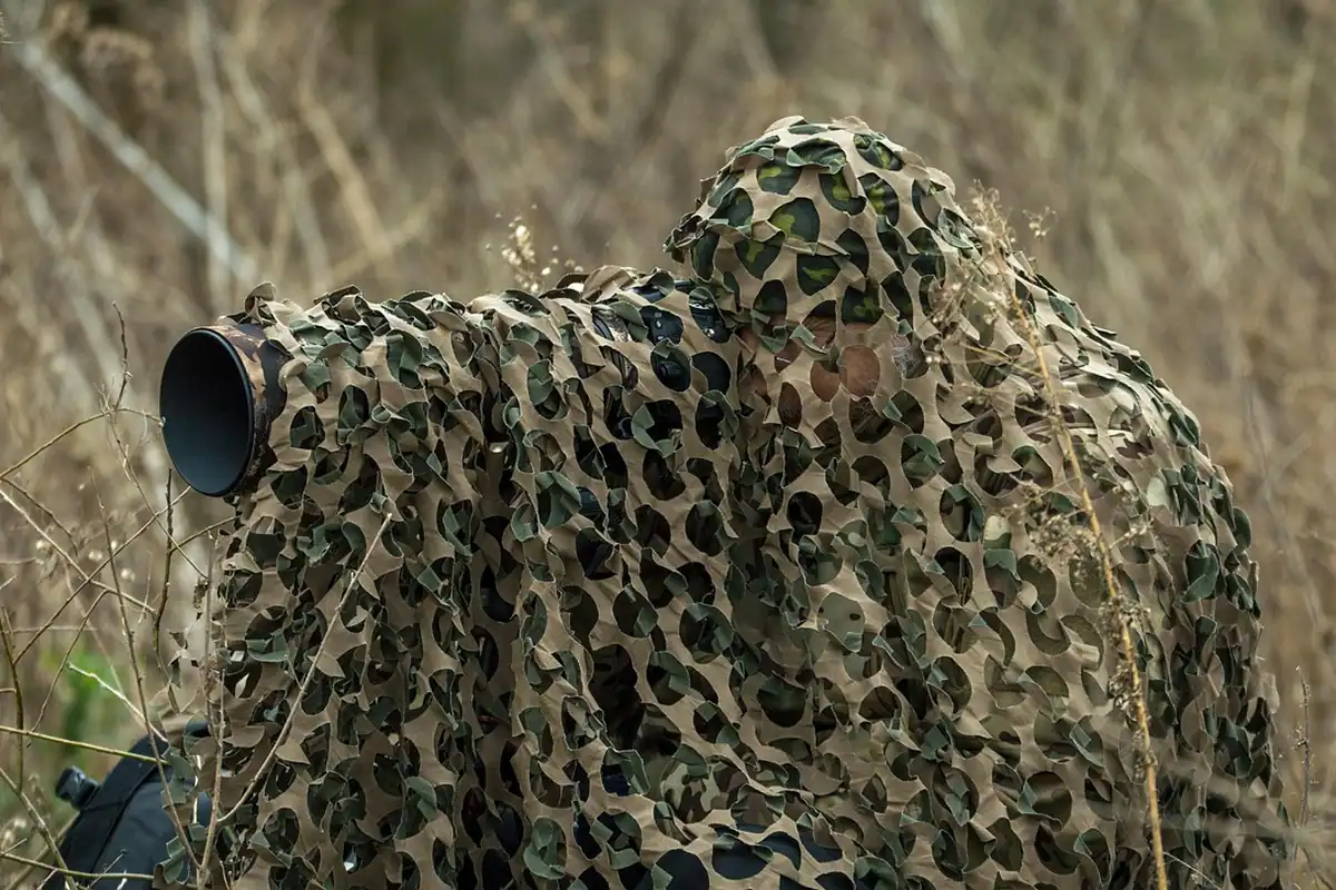 Fotograf camuflat în haine de vânătoare, cu un teleobiectiv lung îndreptat spre iarba înaltă