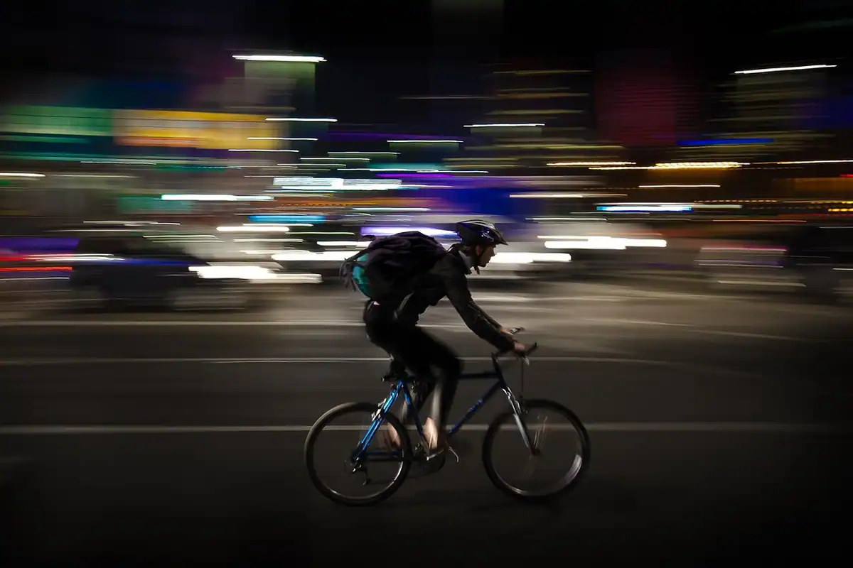 Ciclist pe o stradă noaptea, cu luminile orașului întinse în dâre prin mișcarea aparatului