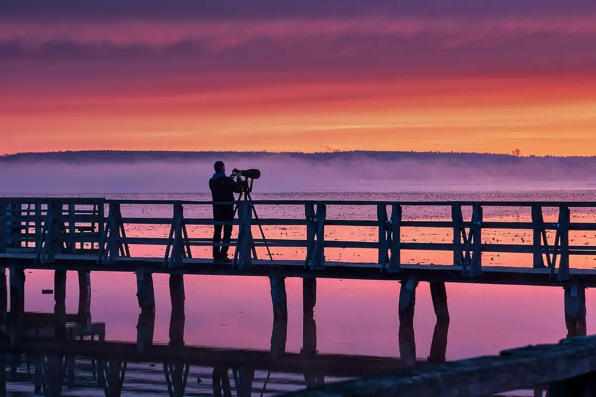 Fotograf cu aparatul pe trepied pe un ponton, la răsărit, cu cerul roz peste apă